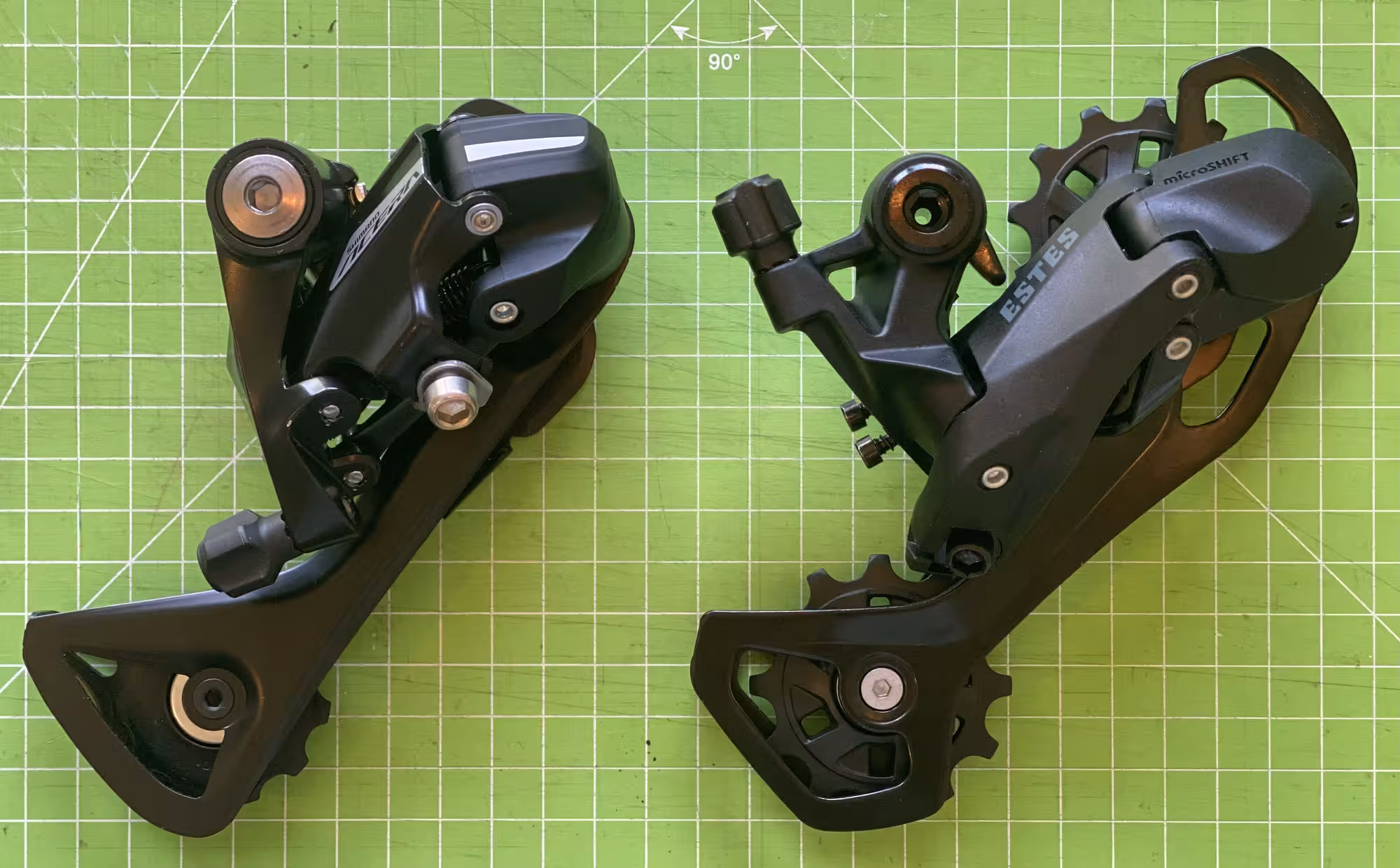 Two rear derailleurs side by side on a green cutting mat. On the left the a little bit smaller Acera, on the right the Estes.